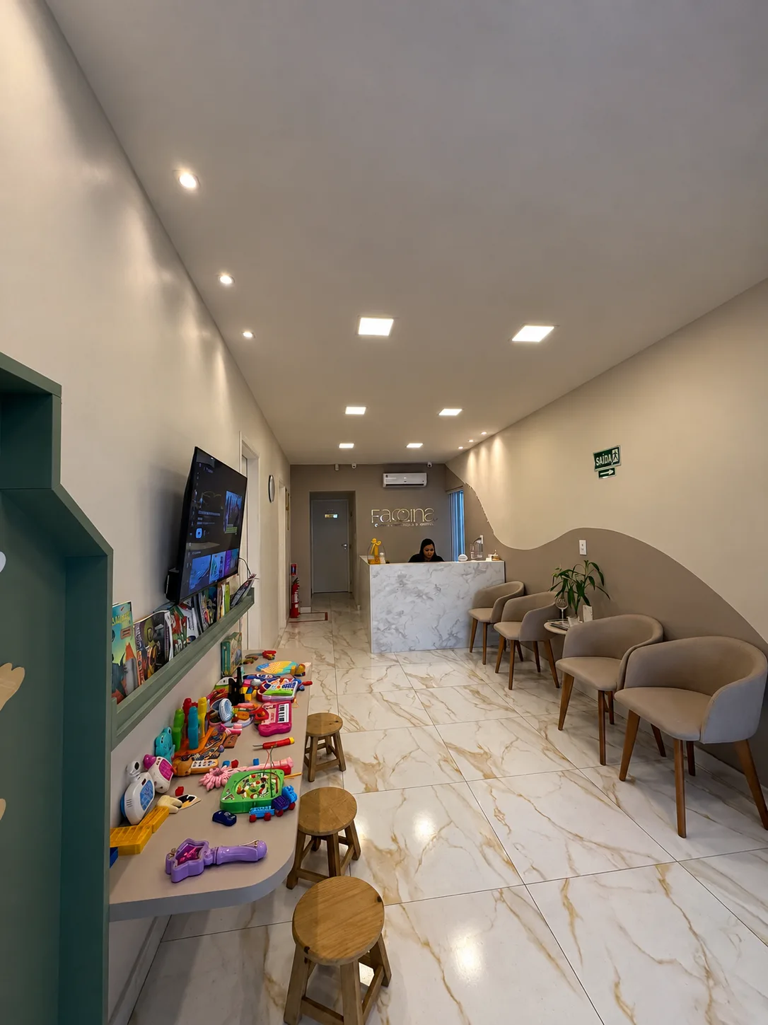 Recepção da Faccina: cantinho kids à esquerda com TV, brinquedos e banquinhos de madeira, poltronas à direita e o balcão ao fundo